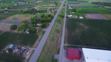 Drone-shot-over-a-well-travelled-road-in-British-Columbia-farm-country,-the-shot-follows-a-car-and-pans-up-to-reveal-the-mountains-in-the-background