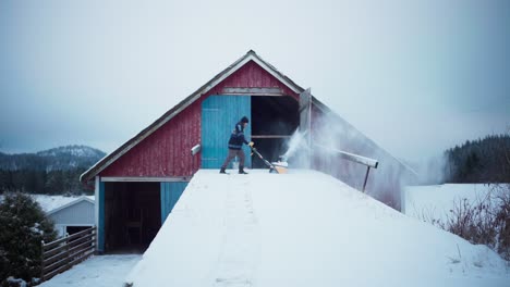 A-Man-With-Electric-Snow-Thrower-Removing-Ice-Near-Wooden-Barn-House