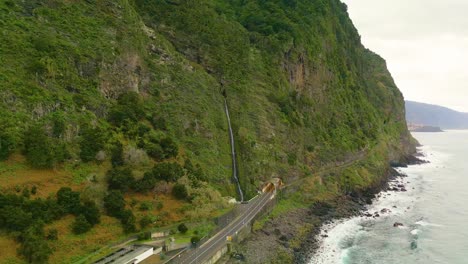 Aerial-view-of-waterfall-on-green-mountains-at-coastal-road-in-Madeira-Island,-Portugal---Atlantic-ocean-on-the-right