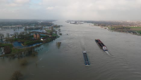 Aerial-view-of-the-historic-Slot-Loevestein-castle-surround-by-flood-waters-as-two-ships-pass-down-the-Waal-River-near-Gorinchem-Netherlands,-after-heavy-rains-pummel-Northern-Europe