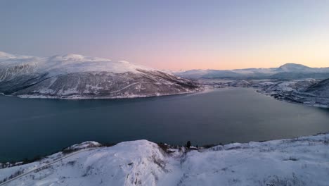 Langsam-Schwenkende-Luftaufnahme-Auf-Dem-Gipfel-Eines-Schneebedeckten-Berges,-Umgeben-Von-Einem-Großen-See-Und-Schneebedeckten-Bergen-Im-Winter-In-Ersfjordvegen,-Norwegen