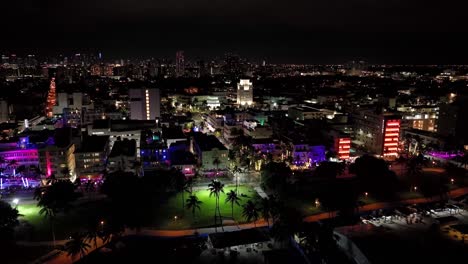 Vibrant-nightly-aerial-drone-footage-of-Miami,-USA:-slow-descent-forward-movement-from-the-seaside,-showing-buildings,-roads,-skyscrapers,-with-many-different-colored-lights