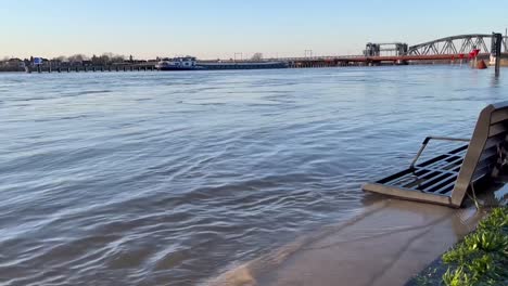 Strong-current-and-calm-waves-reaching-bench-on-the-countenance-boulevard-hit-by-water-during-winter-high-level-of-river-IJssel-showing-partly-flooded-boulevard