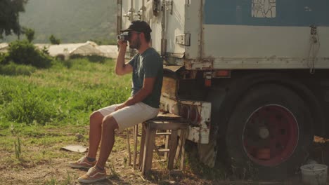 Young-man-sits-on-worn-wooden-bench-behind-semi-truck-raising-vintage-camera-to-face