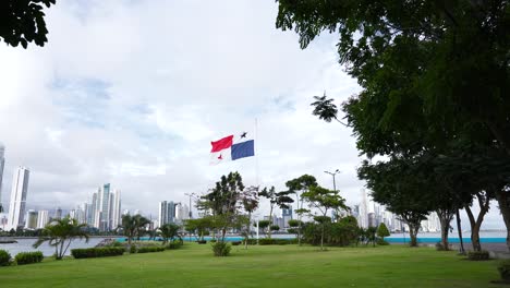 Panama's-national-flag-waving-the-wind,-at-a-park,-against-modern-tall-buildings-and-skyscrapers-near-the-coastline