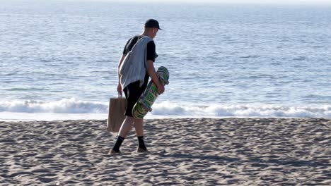 A-skateboarder-carrying-his-board,-a-towel,-and-paper-bag-across-the-beach-on-a-sunny-day