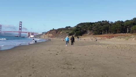 Two-people-walking-in-the-distance,-away-from-the-camera,-along-the-beach-with-the-ocean-and-the-Golden-Gate-bridge-in-the-background