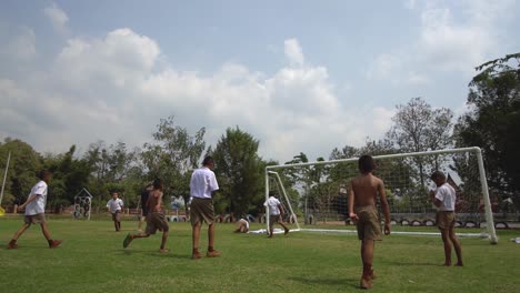 Felices-Niños-De-Escuela-Tailandesa-Jugando-Fútbol-En-Campo-Verde,-Cámara-Lenta-Cinematográfica