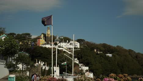 European-Union-Flag-Waving-In-Foreground,-Two-Welsh-Flags-With-Portmeirion-Village-In-Background,-People-Walking