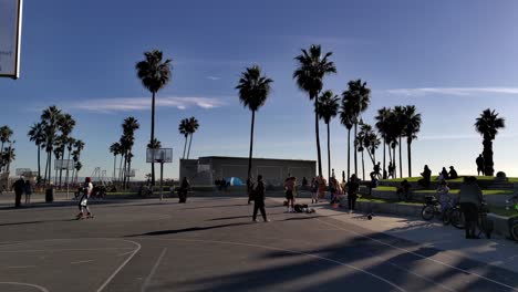 People-hanging-out-and-riding-skateboards-at-the-Venice-Beach-Boardwalk-basketball-courts-in-the-evening-in-Los-Angeles,-California,-USA---Wide-shot
