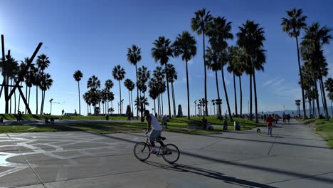 Slow-panning-shot-at-Venice-Beach-Boardwalk-with-people-walking-around-and-biking-past-palm-trees-and-art-sculpture-in-Los-Angeles,-California,-USA