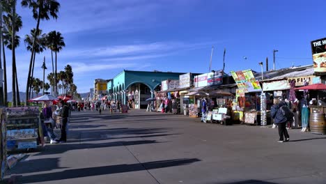 People-walking-in-front-of-stores-at-the-Venice-Boardwalk,-sunny-day,-in-Los-Angeles,-California,-USA---Static-view