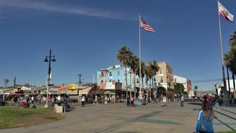 People-walking,-riding-bikes-and-skateboards-at-the-Venice-Beach-Boardwalk-with-the-American-and-Califonia-flag-flying-in-the-background---Static-Shot