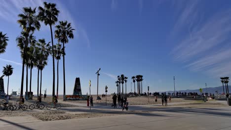 Eine-Gruppe-Von-Menschen,-Die-An-Einem-Sonnigen-Abendtag-In-Los-Angeles,-Kalifornien,-USA,-Mit-Dem-Fahrrad-Den-Radweg-Am-Venice-Beach-Boardwalk-Entlang-Fahren-Und-Im-Hintergrund-Schlittschuh-Laufen