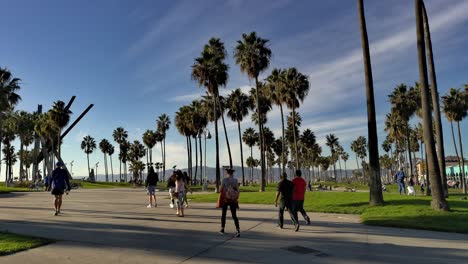 Gente-Caminando-Por-El-Paseo-Marítimo-De-Venice-Beach-Por-La-Tarde-Con-Palmeras-Y-Esculturas-De-Arte-Al-Fondo-En-Los-Ángeles,-California,-EE.UU.---Toma-Amplia