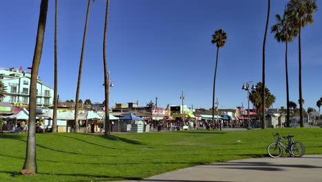 Menschen,-Die-An-Der-Strandpromenade-Von-Venice-Entlang-Des-Strips-Im-Hintergrund-Mit-Geschäften-Und-Ständen-An-Einem-Sonnigen-Nachmittag-In-Los-Angeles,-Kalifornien,-USA,-Spazieren-–-Statische-Handaufnahme