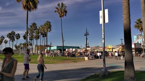 People-walking-and-riding-bikes-on-a-sunny-afternoon-at-the-Venice-Beach-boardwalk-with-grassy-park-and-palm-trees-in-the-background-in-Los-Angeles,-Califonia,-USA