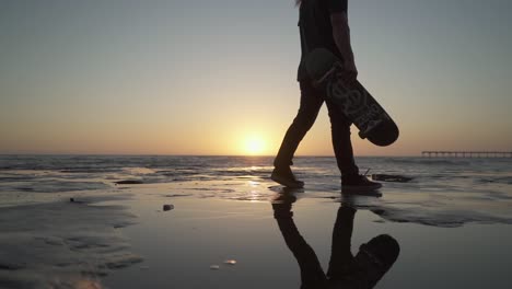 Cinematic-slow-motion-shot-of-young-male-with-skateboard-walking-on-cliffs-near-the-ocean-during-sunset