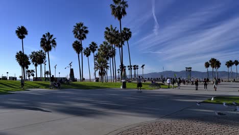 Menschen,-Die-Während-Covid-19-Auf-Skateboards-Auf-Der-überfüllten-Strandpromenade-Von-Venice-Beach-Vorbeikommen,-Mit-Skatepark-Und-Kunstskulpturen-Im-Hintergrund-In-Los-Angeles,-Kalifornien,-USA