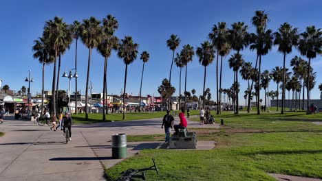 Blick-Auf-Die-Promenade-Von-Venice-Beach-Mit-Menschen,-Die-Mit-Der-Amerikanischen-Und-Kalifornischen-Flagge-In-Los-Angeles,-Kalifornien,-USA-Spazieren-Gehen-Und-Fahrrad-Fahren-–-Schwenkaufnahme