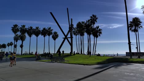 Wide-shot-on-Venice-Beach-Boardwalk-across-artwork-and-Art-sculpture-on-a-grassy-lawn-with-people-walking-around-and-dogs-in-Los-Angeles,-California,-USA---Wide-Panning-shots