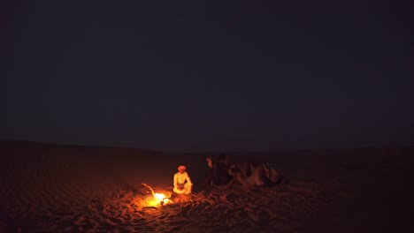 Dusk-in-gloomy-Oman-desert-lit-by-campfire-of-man-with-camel-caravan---Wide-Static-shot