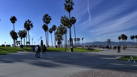 Gente-Caminando-Por-El-Concurrido-Paseo-Marítimo-De-Venice-Beach-Durante-El-Covid-19-Con-Skatepark-Y-Esculturas-De-Arte-Al-Fondo-En-Los-ángeles,-Califonia,-Estados-Unidos