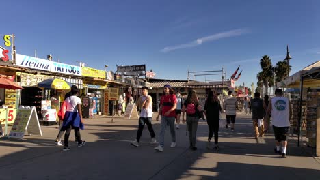 Walking-through-a-crowd-of-people-at-Venice-Beach-boardwalk-with-shops-and-stalls-on-a-sunny-afternoon-in-Los-Angeles,-California,-USA---Walking-shot