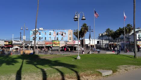 Venice-Beach-Boardwalk-with-people-shopping-around-and-eating-at-restaurants,-sunny-evening,-in-Los-Angeles,-California,-USA---Pan-shot
