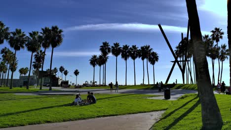 Gente-Relajándose-En-Un-Césped-En-El-Paseo-Marítimo-De-Venice-Beach-Con-Una-Escultura-De-Arte-Gigante-Al-Fondo-En-Una-Tarde-Soleada-En-Los-Ángeles,-California,-Estados-Unidos