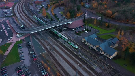 Aerial-view-of-a-VR-train-leaving-a-small-city-station,-gloomy-autumn-day,-in-Finland---circling,-drone-shot