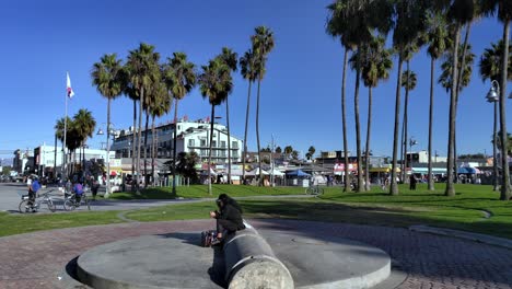 Panning-across-Venice-Beach-Boardwalk-center-looking-at-people-walking-around-and-grassy-parks-with-shopping-center-and-American-and-California-flags-in-the-background-Los-Angeles,-Califonia-USA