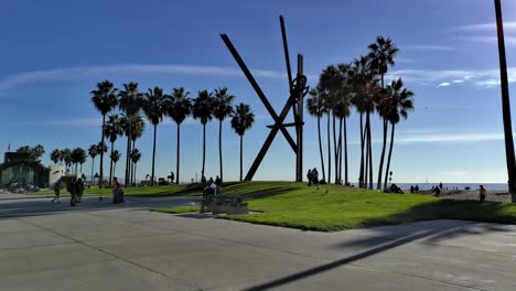 People-walking-around-the-grassy-park-with-giant-art-sculpture-at-Venice-Beach-Boardwalk-on-a-sunny-afternoon-in-Los-Angeles,-California,-USA---Handheld-Static