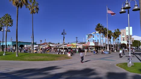 Venice-Beach-Boardwalk-in-the-afternoon-with-people-walking-around-and-riding-bikes-shopping-and-American-flag-and-California-Flag-flying-in-the-background-in-Los-Angeles,-California,-USA---Handheld