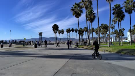 Gente-Andando-En-Bicicleta-Y-Haciendo-Trucos-En-Una-Barandilla-En-El-Paseo-Marítimo-De-Venice-Beach-Con-Un-Skatepark-Al-Fondo-En-Los-Ángeles,-California,-EE.UU.