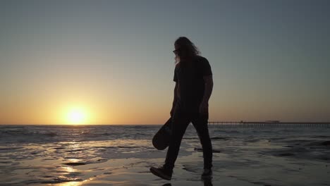 Filmische-Zeitlupenaufnahme-Eines-Jungen-Mannes,-Der-Während-Des-Sonnenuntergangs-Ein-Skateboard-Auf-Klippen-Am-Meer-Hält