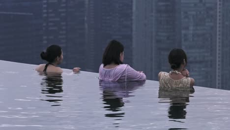 Mujeres-Turistas-Contemplando-El-Paisaje-Urbano-Desde-El-Borde-De-La-Piscina-Infinita-En-El-Hotel-Marina-Bay-Sands,-Singapur---Plano-Medio-Largo