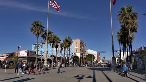 Menschen,-Die-An-Der-Promenade-Von-Venice-Beach-Spazieren,-Mit-Wehenden-Amerikanischen-Flaggen-Im-Wind-Und-öffentlichem-Kunstgebäude-„Touch-Of-Venice“-Im-Hintergrund-In-Los-Angeles,-Kalifornien,-USA