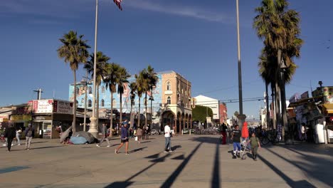Gente-Caminando-En-El-Paseo-Marítimo-De-Venice-Beach,-Tiendas-Y-Puestos-Y-Palmeras-Y-Parques-Cubiertos-De-Hierba-En-El-Fondo-En-Los-Ángeles,-California,-EE.UU.