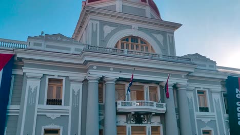 A-man-is-walking-by-in-front-of-a-government-building-that-has-a-big-flag-of-Cuba-and-other-flags-in-the-main-plaza-of-the-city-of-Cienfuegos,-Cuba