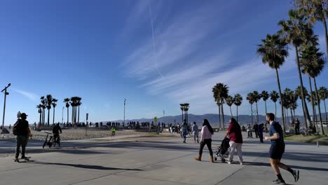 People-walking-around-Venice-Beach-Boardwalk-with-bikes-and-strollers-enjoying-a-sunny-afternoon-in-Los-Angeles,-California,-USA---Panning-shot