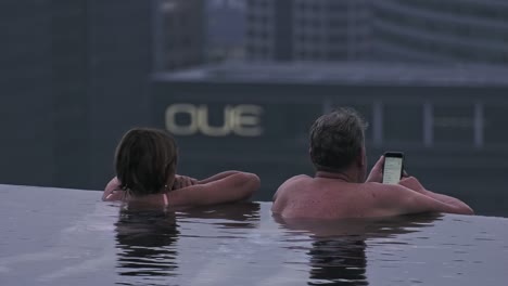 Backwards-facing-middle-age-couple-resting-on-edge-of-infinity-pool-at-Marina-Bay-Sands,-Singapore---Long-Medium-Static-shot