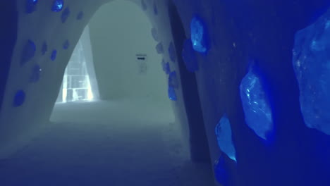 Lumps-of-Glowing-Blue-Ice-Wall-Lights-in-Cavernous-Corridor-at-Snow-Village-Lapland,-Panning-Shot