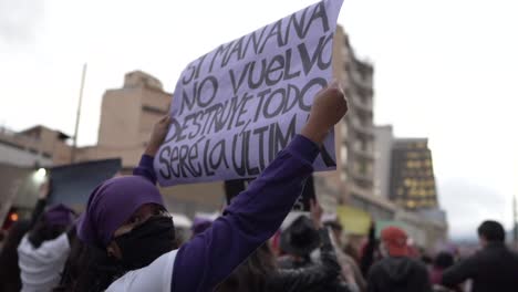 A-woman-wearing-a-purple-handkerchief-holds-up-a-sign-during-the-International-Women's-day-in-Quito,-Ecuador,-She-is-wearing-a-mask-mask-due-to-covid-19