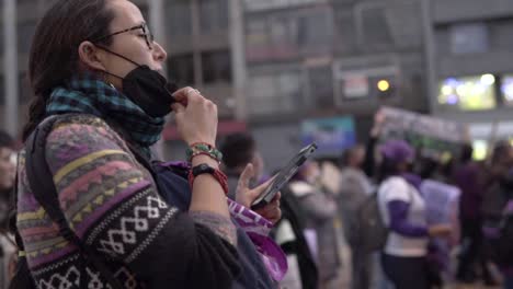 Eine-Frau-Trägt-Eine-Schwarze-Maske-Und-Beobachtet-Ihr-Mobiltelefon-Während-Einer-Protestaktion-Am-Internationalen-Frauentag-In-Quito,-Ecuador,-Menschen-Auf-Dem-Rücken-Singen-Und-Halten-Schilder-Hoch