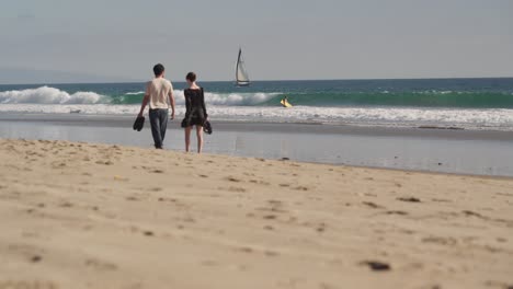 Back-View-of-Straight-Couple-Walking-Barefoot-in-Sand-of-Santa-Monica-Beach-in-Front-of-Ocean-Waves-and-Sailboat