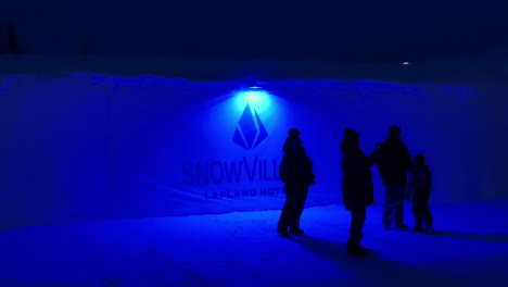 Gente-Esperando-Afuera-Por-La-Noche-Junto-Al-Letrero-De-Entrada-Iluminado-Para-El-Hotel-Snow-Village-Laponia