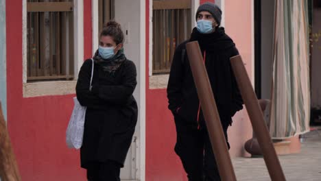 Pareja-Joven-Caminando-Con-Máscaras-Protectoras-Azules-En-La-Isla-De-Burano,-Venecia.
