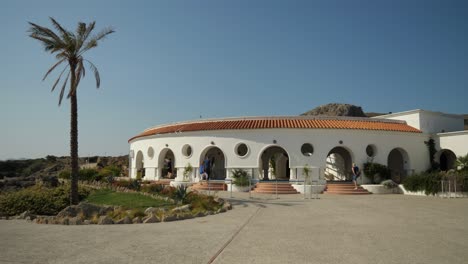Eye-level-view-of-a-round-building-at-Kallithea-Springs-with-palm-tree-and-clear-sky,-People-walking