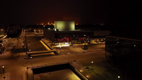 Aerial-view-ascending-in-front-of-the-illuminated-Oulu-Theatre,-winter-night-in-Finland